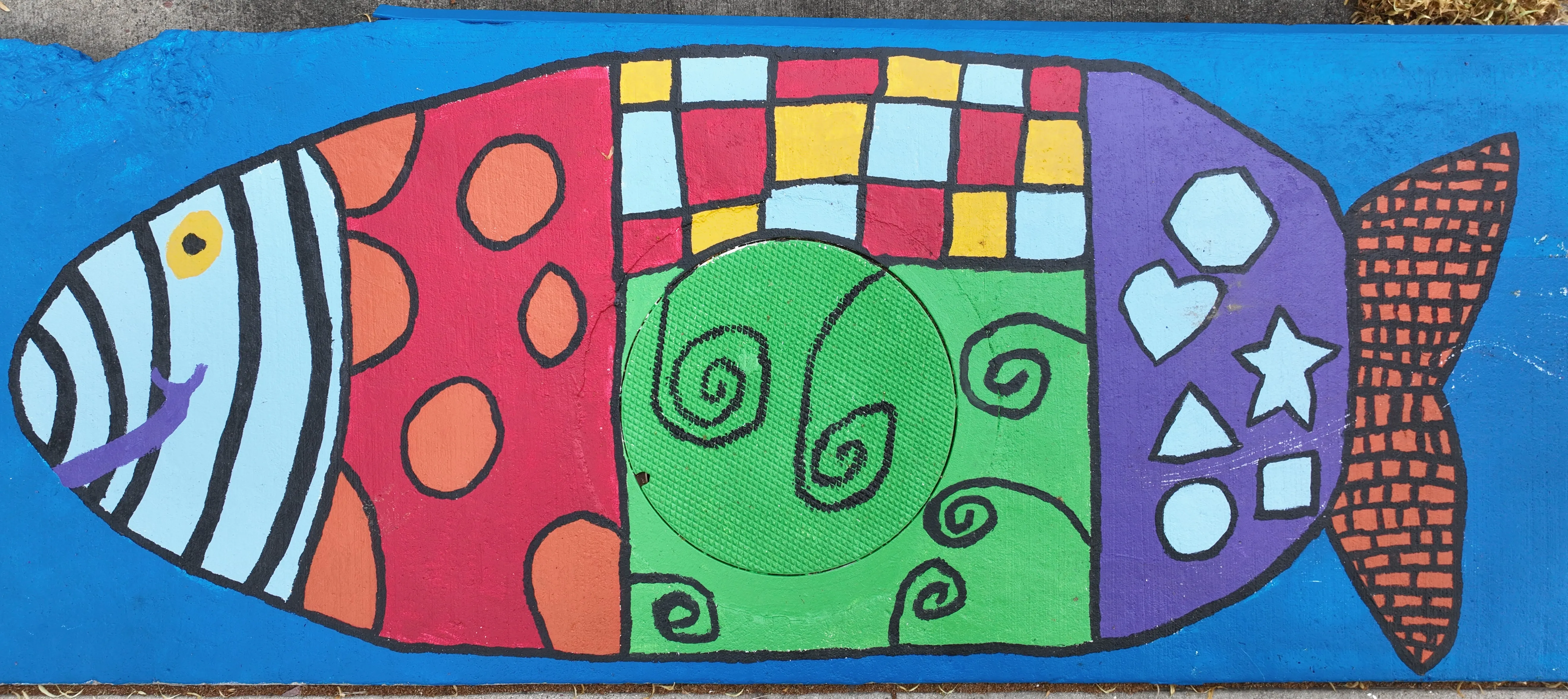 A mural on a storm inlet drain depicting a simple two dimension shape with segments decorated with strips, dots, and other shapes.