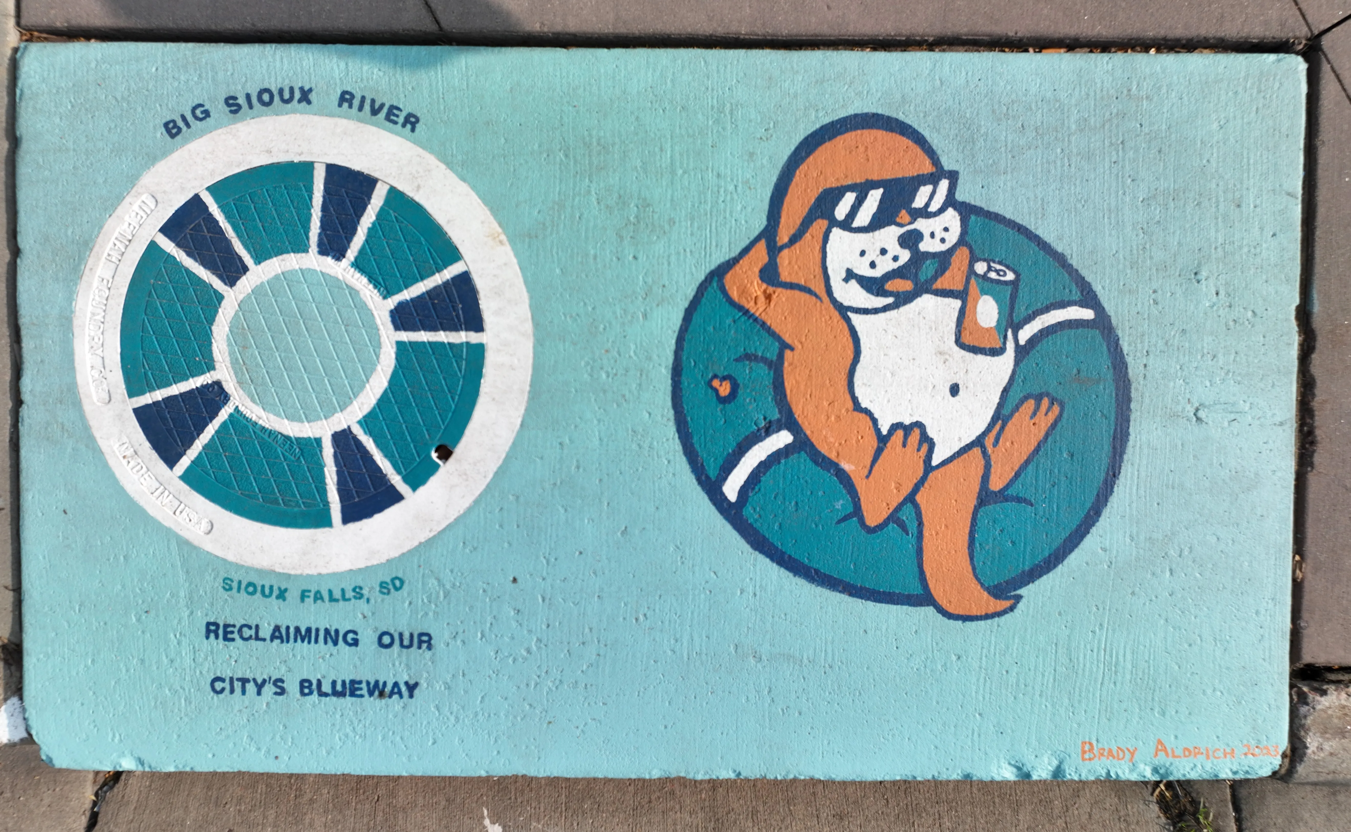 A mural on a storm inlet drain featuring an otter with sunglasses and a beverage relaxing in a pool tube. Text reads "Big Sioux River, Sioux Falls SD - Reclaiming our City's Blueway"