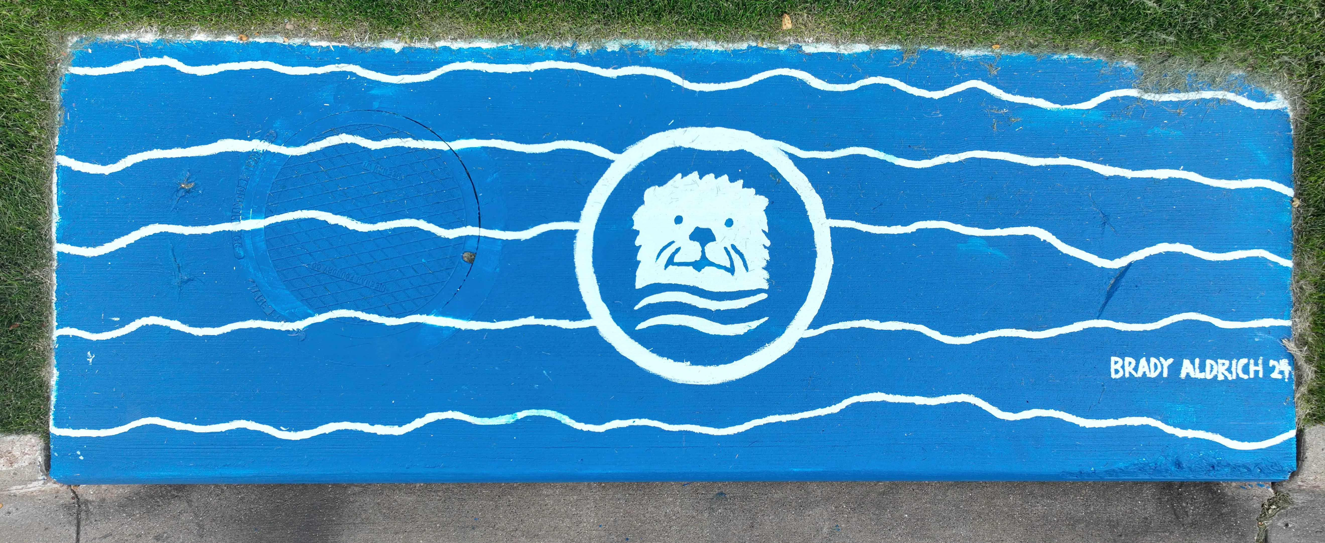 A mural on a storm inlet drain featuring a blue background and blue wavy lines. Centered is an otter's face.