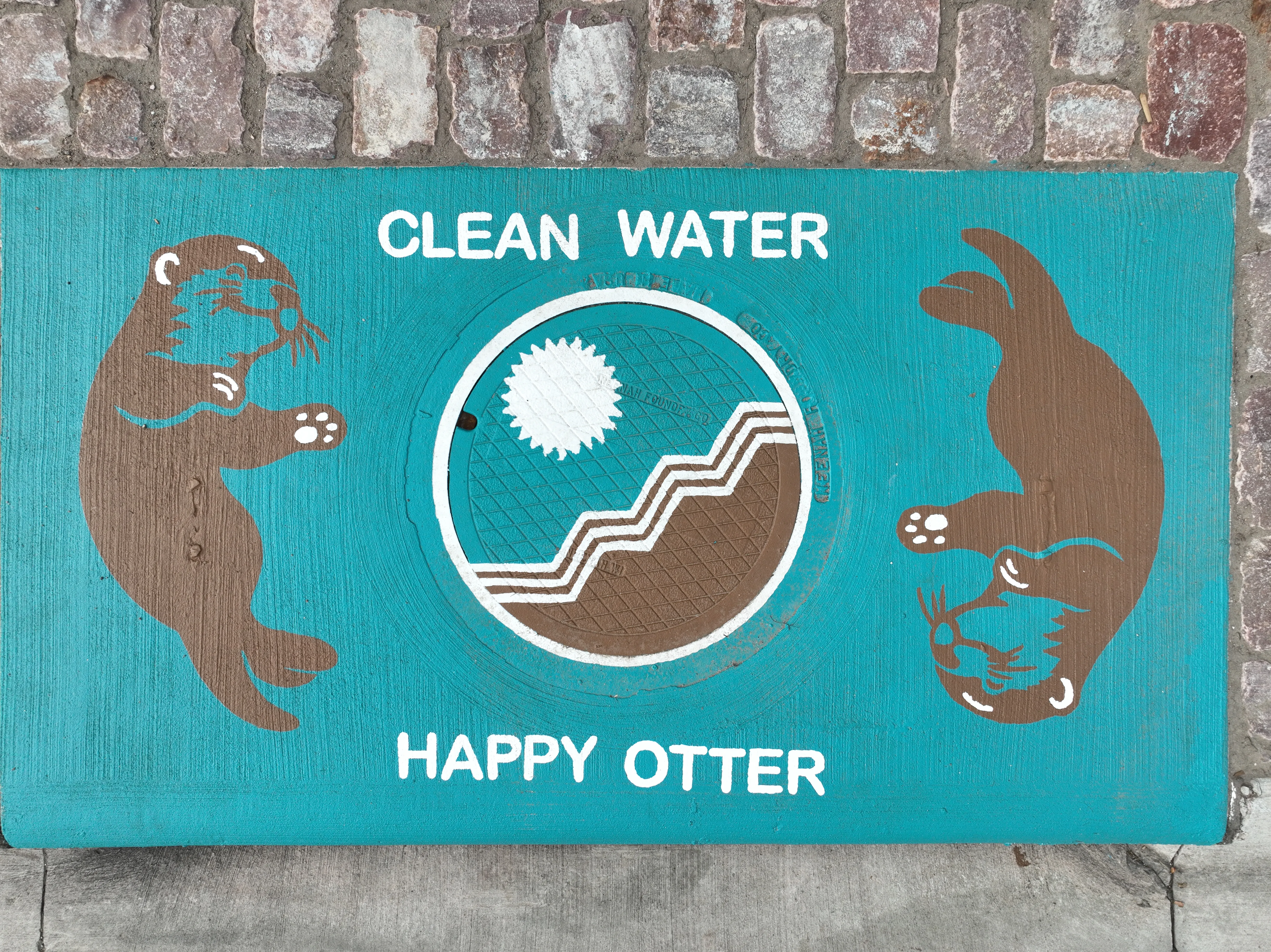 A storm drain mural featuring two otters facing on either side facing inwards towards a City of Sioux Falls design on the manhole cover, with a blue background.