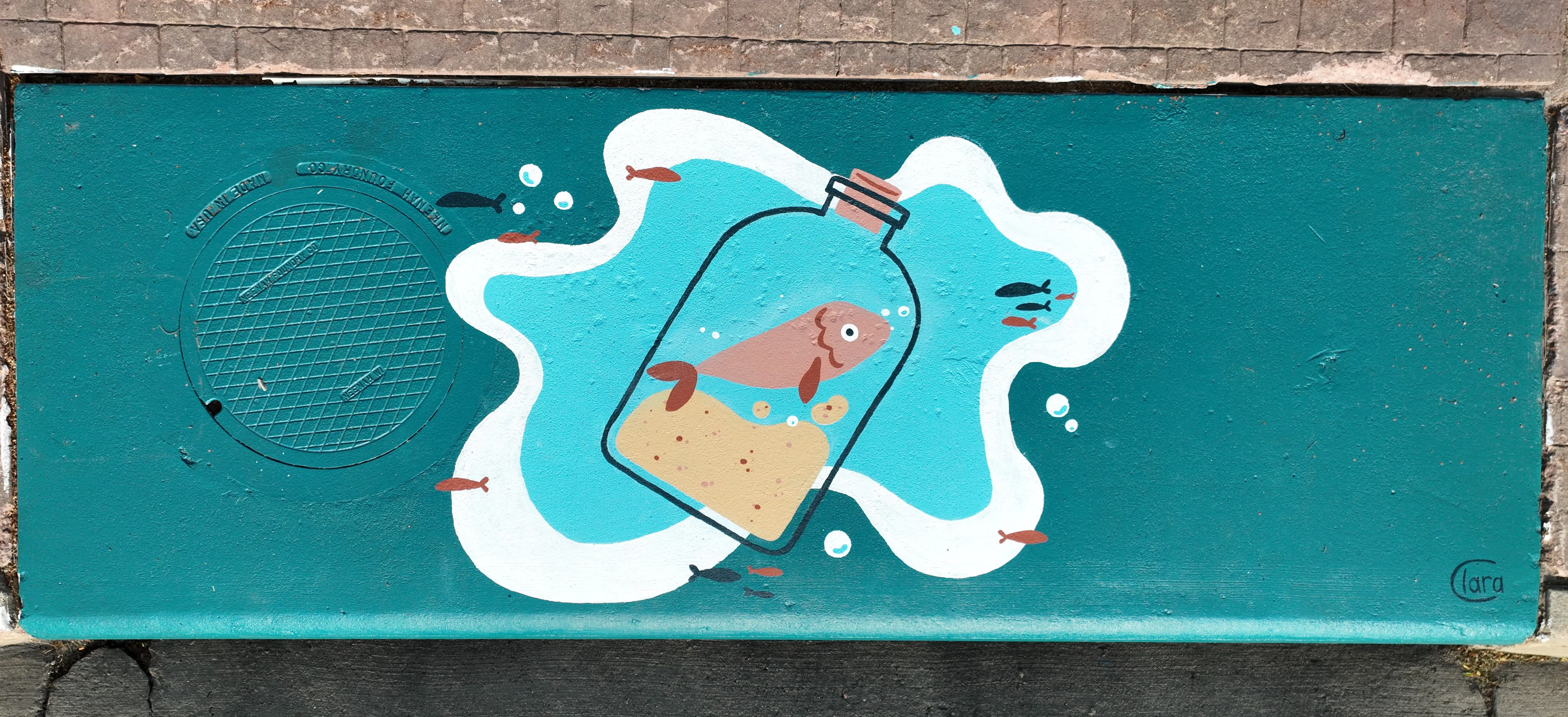 A mural on a storm inlet drain featuring a fish inside a bottle and other smaller fish swimming around.
