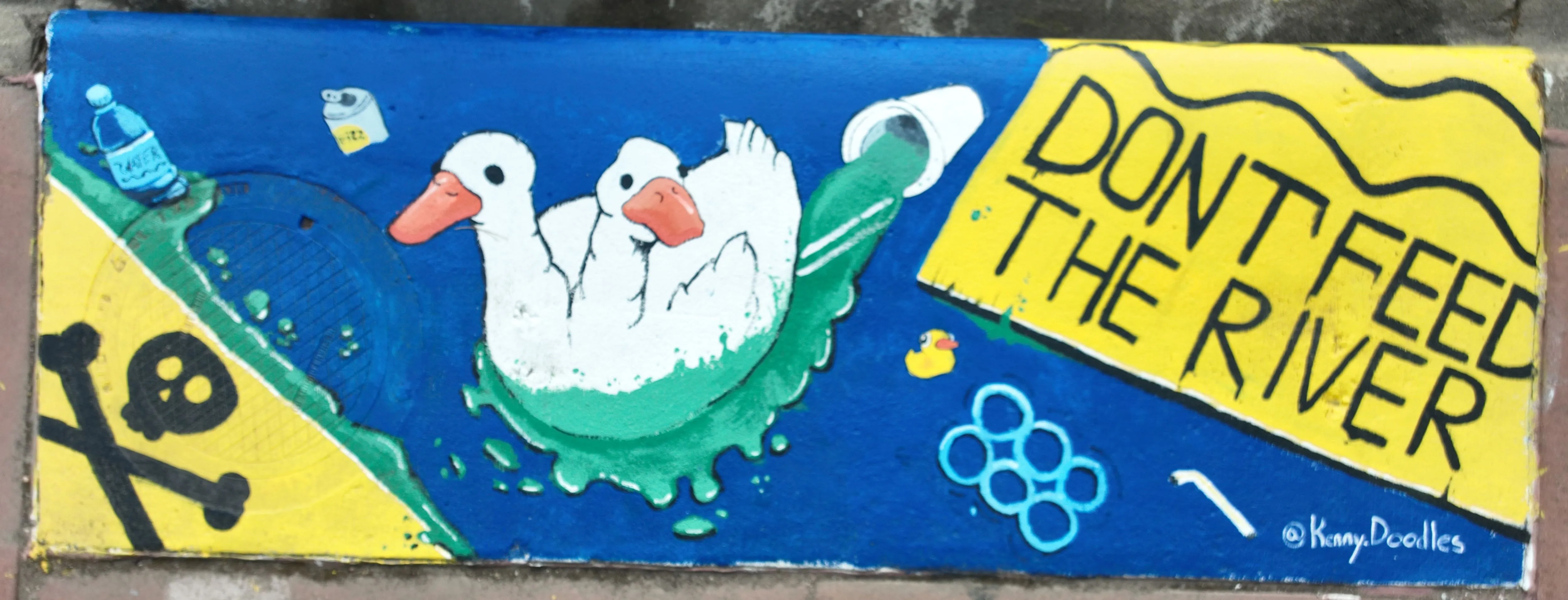 A mural on a storm inlet featuring a mutant two-headed duck in the center surrounded by trash and green sludge. Text reads "Don't feed the river"