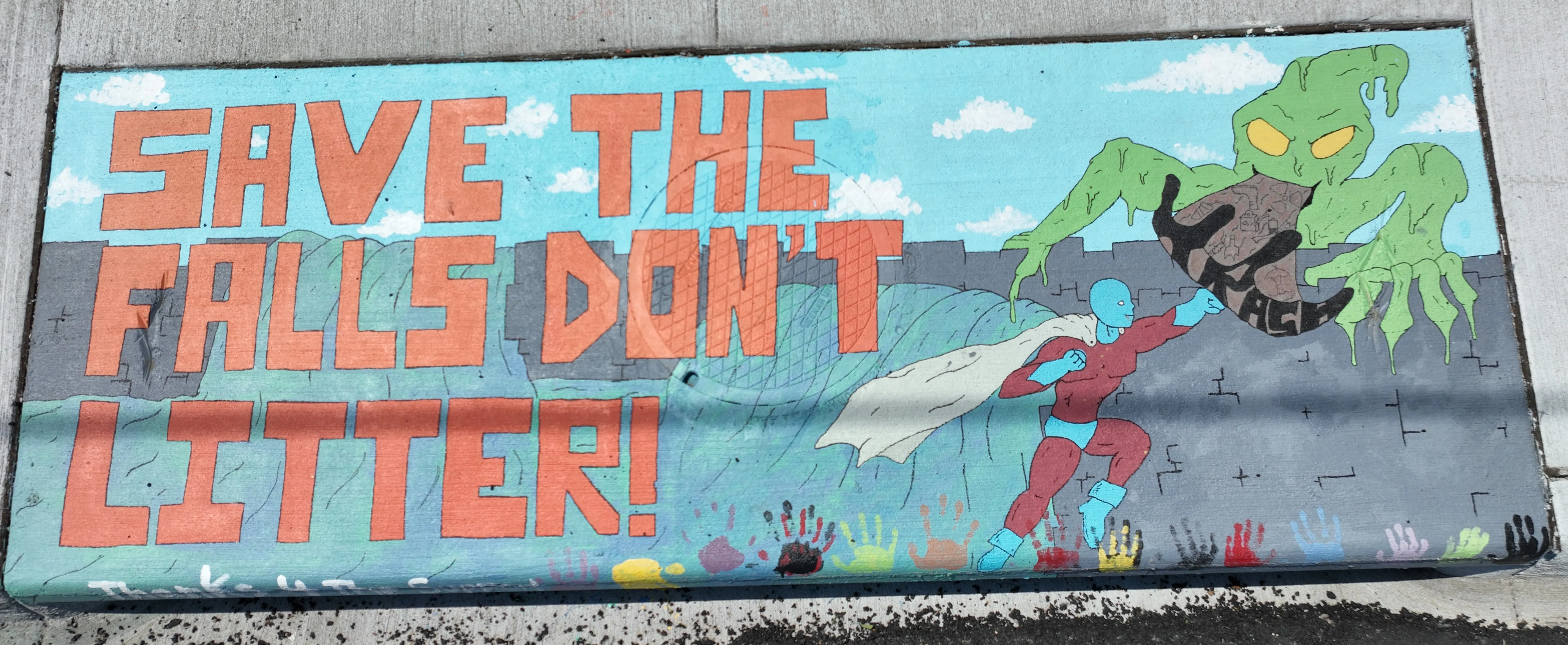 A mural on a storm inlet drain featuring a superhero fighting a green monster spewing a gray substance labeled "Trash". Large cartoon block letters on the left read "Save the Falls Don't Litter!"